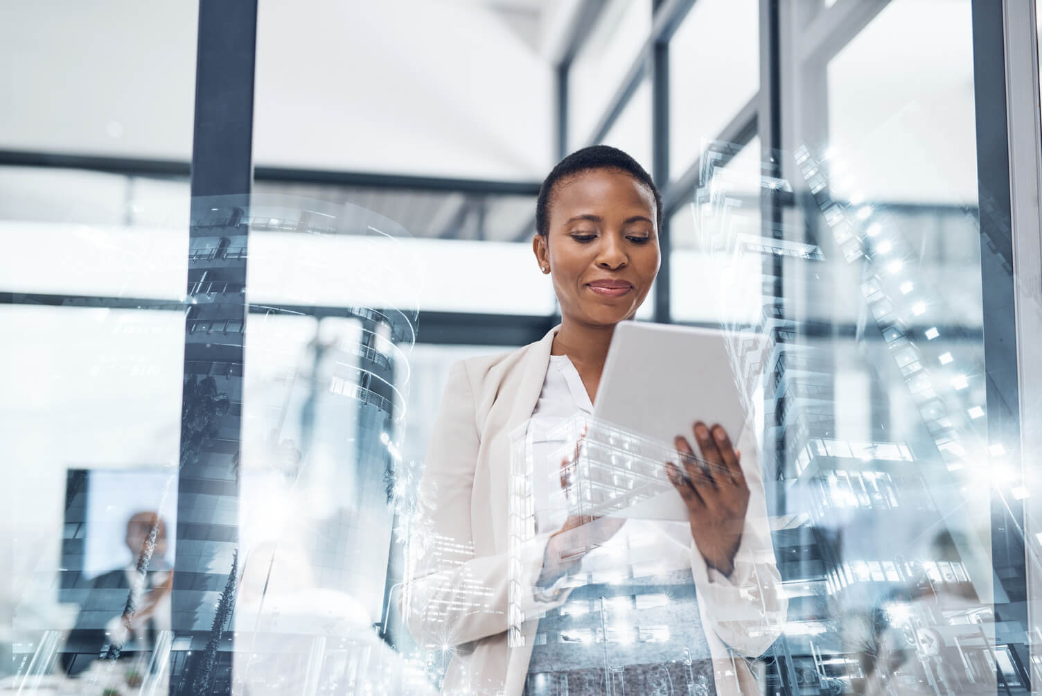 Person standing in a modern office, holding a tablet, with reflections of digital graphics on glass walls.