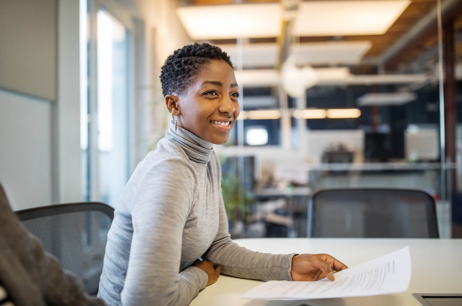 A Black woman in a gray turtleneck sits at a desk holding documents in a bright, modern office workspace.