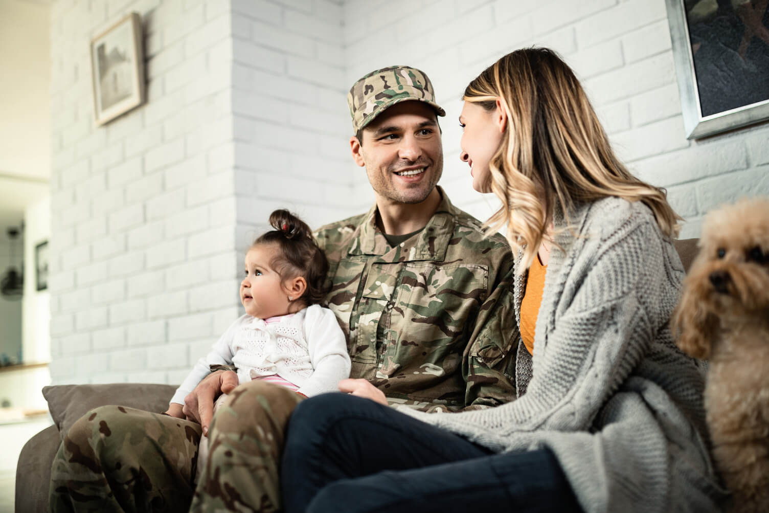 Person in military uniform sitting on a couch with a child on their lap and another adult beside them, in a cozy living room with a white brick wall and a small dog nearby.