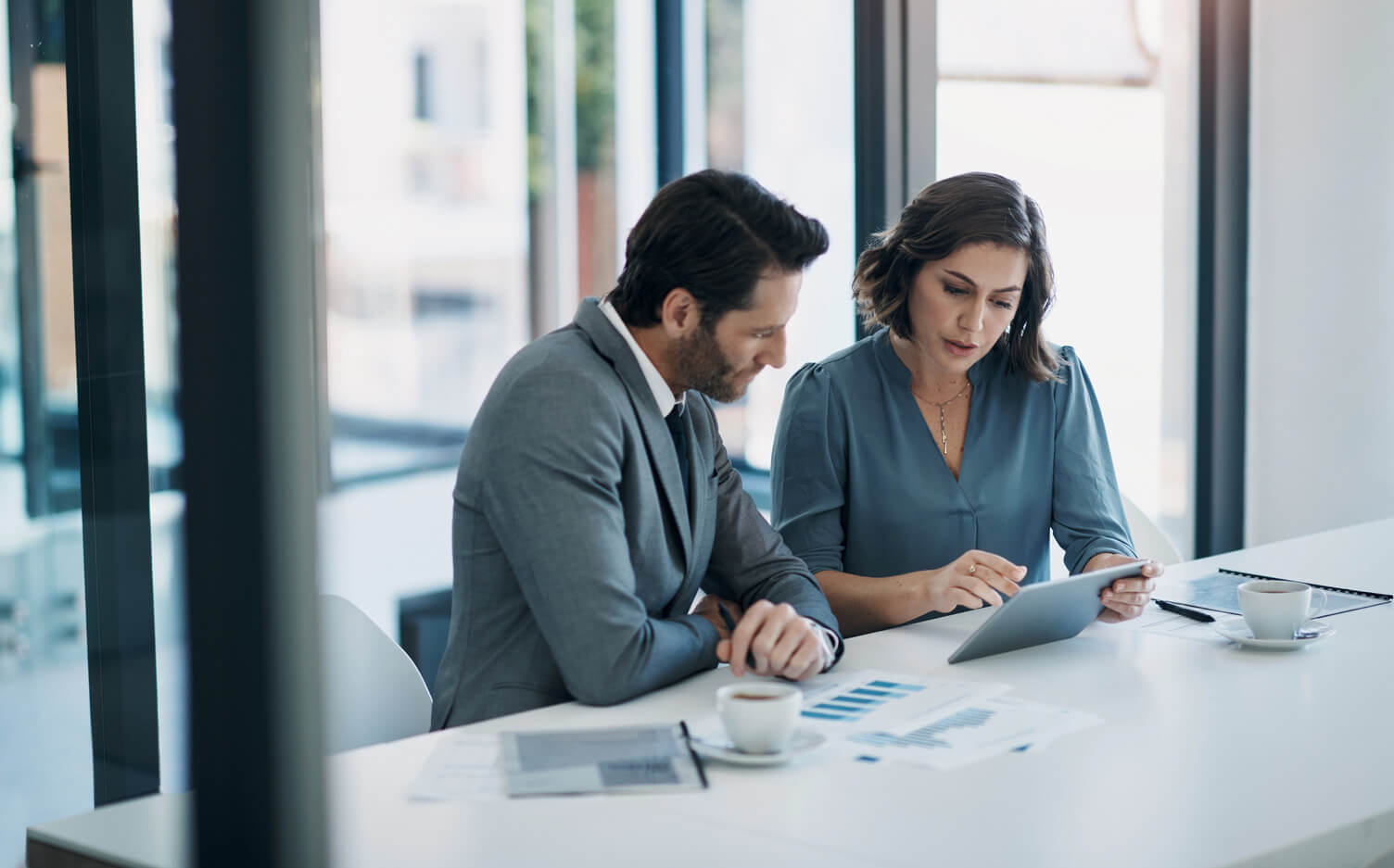 Two business professionals sit at a conference table reviewing information on a tablet, with printed charts and coffee cups in front of them.
