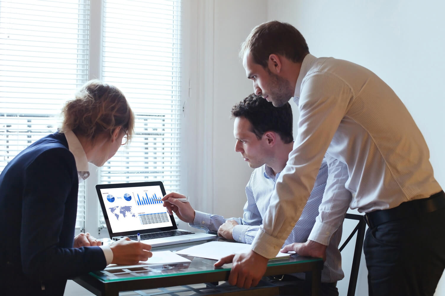 Three adults in business attire sit at a desk table in a modern office, reviewing capital markets data on laptops