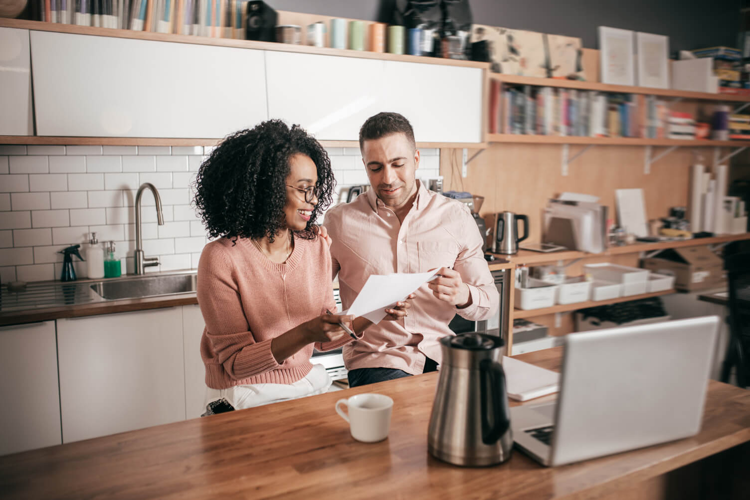 Black woman and light-skinned man review printed documents at a kitchen table with laptop and coffee, suggesting employment verification.