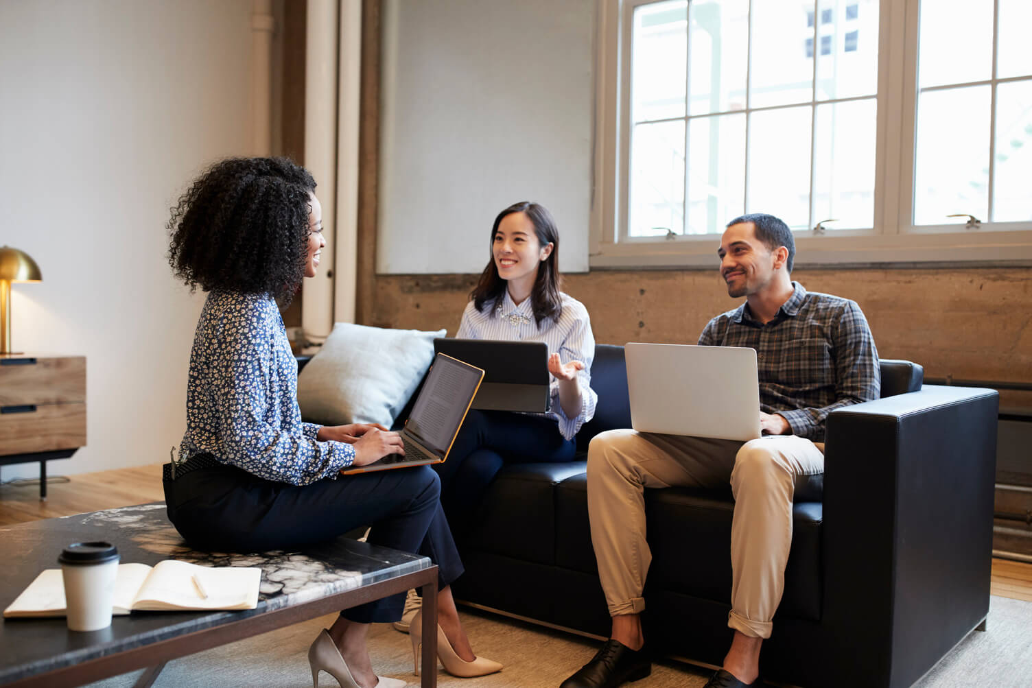 Black woman and two colleagues—a woman and a man—sit on a couch with laptops and documents, discussing information in a bright office setting.