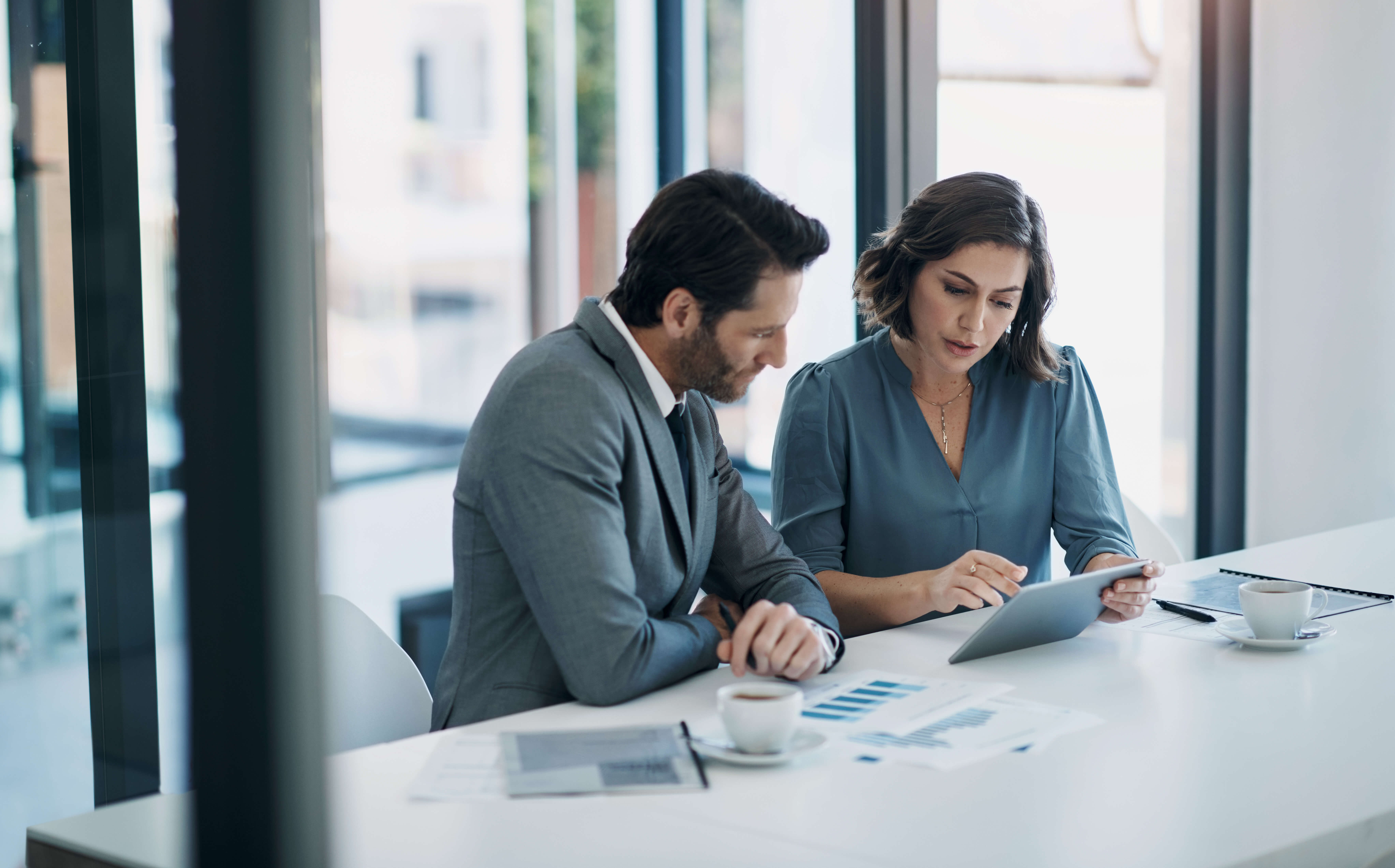 Two business professionals sit at a conference table reviewing information on a tablet, with printed charts and coffee cups in front of them.