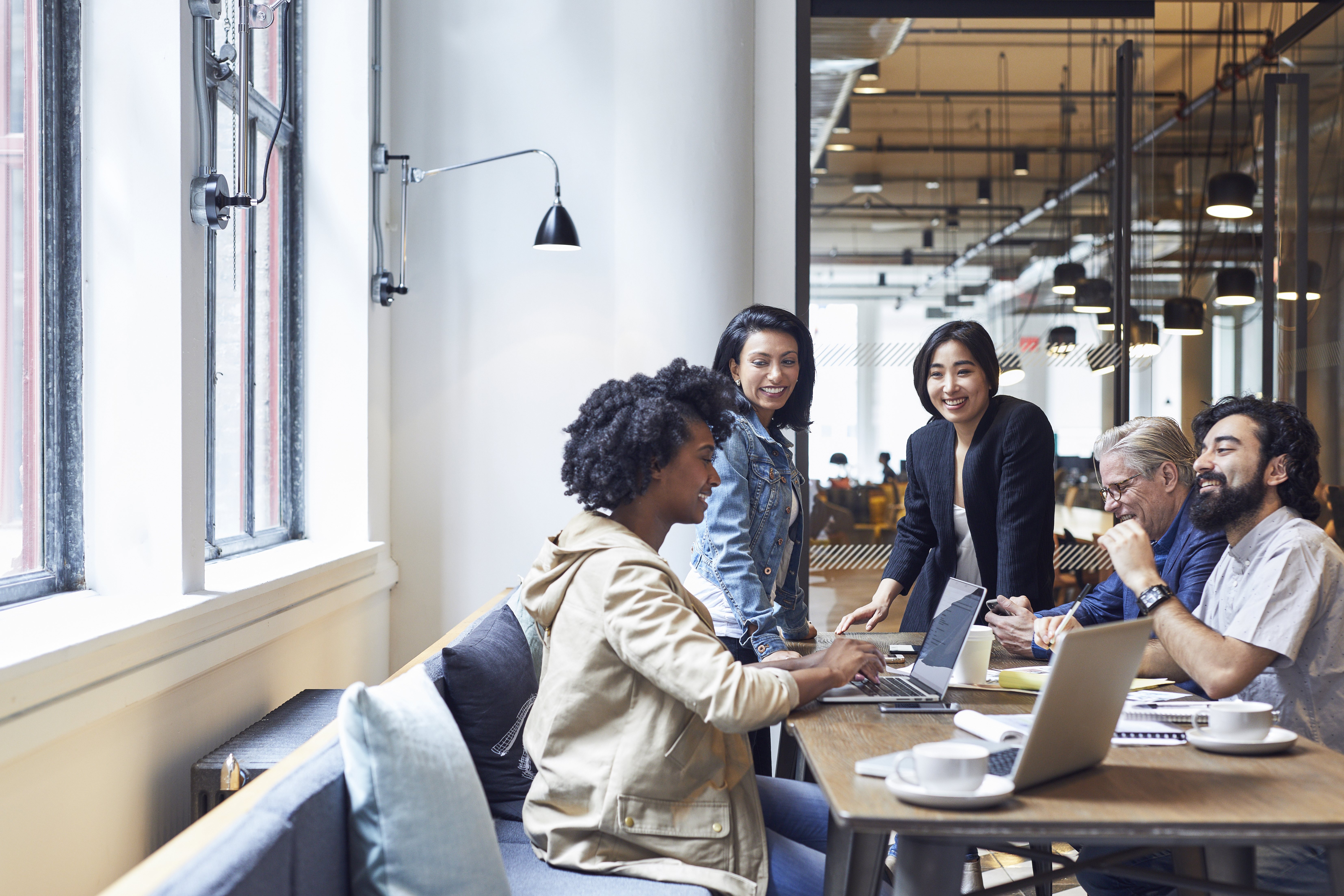 Diverse group of adults collaborate at a long table in a modern office, using laptops and documents in a bright, open workspace.